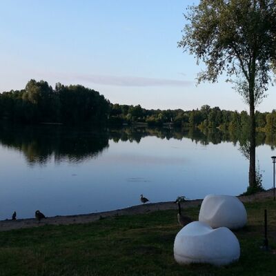 Blick auf den Hegbachsee, im Vordergrund Wiese, zwei weiße Sitzkiesel und ein Baum, Abendämmerung