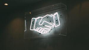 Bild vergrößern: Lampe aus weißen Neonröhren, die zwei in einandergeschlungene Hände bilden. Schwarzer Untergrund