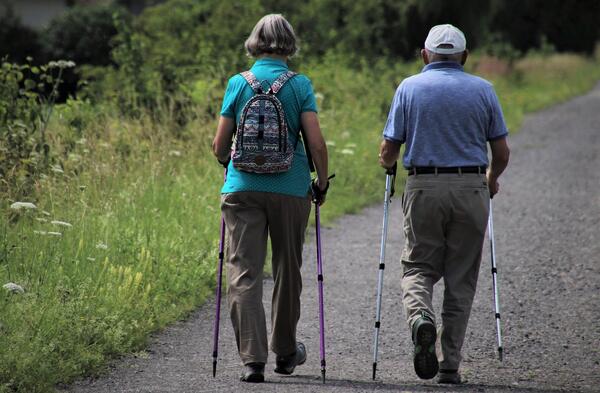 Bildansicht zeigt zwei ältere Menschen von hinten, links eine Frau mit Rucksack und recht einen Mann mit Kappe, die beiden mit Walkingstöcen auf einem Schotterweg laufen, links eine grüne natürlich bewachsene Wiese