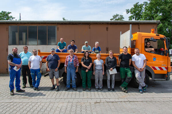 Bild vergrößern: Gruppenfoto der Beschäftigten des Bauhofs, teilweise stehen die Mitarbeiter vor einem orangefarbenen kleinen LKW mit Ladefläche, teilweise auf der Ladefläche, im Hintergrund eine Halle des Bauhofs, blauer Himmel