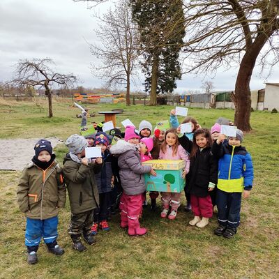 Winterlich gekleidete Kinderschar steht um bunt bemalten Briefkasten aus Pappe, auf dem "KiJuPa" steht. Einige der Kinder strecken bemalte Postkarten in die Höhe, Szene ist auf dem Außengelände der Nauheimer Natur-Kita auf einer grünen Wiese, kahle Bäume im Hintergrund