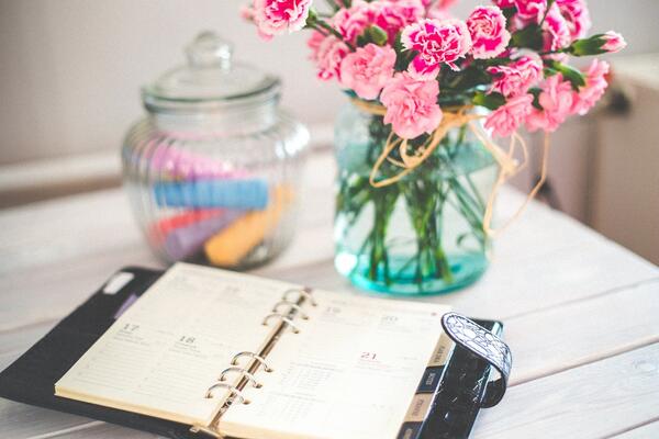 Bild vergrößern: Auf einem weißen Holztisch liegt ein aufgeschlagener Terminkalender, im Hintergrund eine Glasvase, in der rosafarbene Blumen stehen sowie ein weiteres Glasbehältnis mit Glasdeckel, in dem bunte Gegenstände liegen